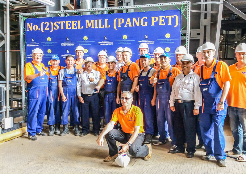 На изображении представлена группа рабочих в спецодежде и защитных касках, стоящих перед баннером с надписью «No.(2) STEEL MILL (PANG PET)».